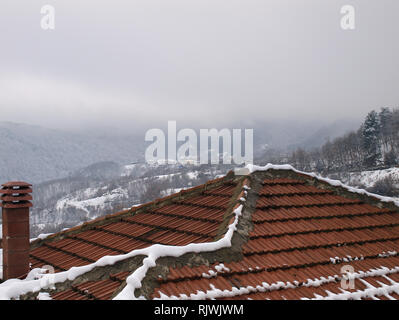 Tegole rosse del tetto e del camino di una casa di villaggio con montagne coperte di neve e cielo molto nuvoloso in background Foto Stock