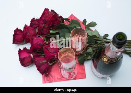 Le rose red che giace piatto sul tavolo bianco all'esterno insieme con un aprire una bottiglia di champagne o vino frizzante e due bicchieri riempiti pronto per la celebrazione Foto Stock