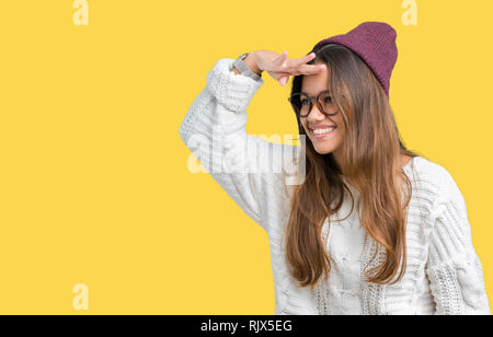 Giovane bella bruna hipster donna che indossa occhiali e cappello invernale su sfondo isolato molto felice e sorridente guardando lontano con la mano sopra Foto Stock