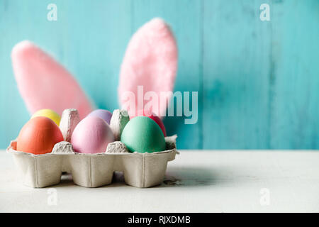 Color pastello di uova di pasqua in una scatola per uova e orecchie di coniglio dietro con un legno sfondo turchese Foto Stock