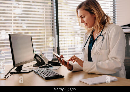 Medico femmina seduto alla sua scrivania e lavorando al suo digitale compressa. Medico usando un computer tablet nel suo ufficio. Foto Stock