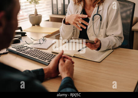 Medico donna parlando del trattamento con paziente di sesso maschile al suo clinic desk. Ritagliato colpo di medico femmina tenendo una tavoletta digitale parlando con Foto Stock