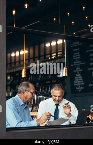 Ripresa verticale di due uomini anziani che si incontrano in un pub e discutono su un documento. uomini d'affari seduti vicino a una finestra al bar e che parlano con un rivenditore Foto Stock