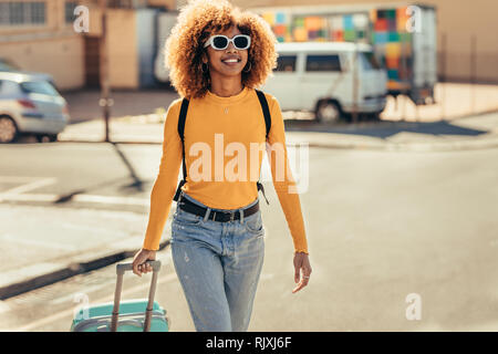 Donna afro american viaggiatore in vacanza a piedi intorno alla città tirando la sua valigia trolley. Sorridente turista donna che indossa gli occhiali da sole e uno zaino wa Foto Stock