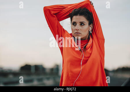 Adatta la giovane donna in tuta in piedi all'aperto e allungando le braccia. donna in forma fisica con auricolari in pausa dopo una corsa mattutina. Foto Stock