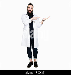 Medico con capelli lunghi che indossa medical e il cappotto di uno stetoscopio stupiti e sorridente alla fotocamera durante la presentazione con la mano e puntando con il dito. Foto Stock