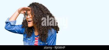 Giovane bella donna con capelli ricci con gli occhiali molto felice e sorridente guardando lontano con la mano sopra la testa. Concetto di ricerca. Foto Stock