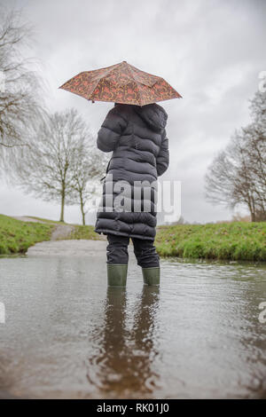 Kidderminster, Regno Unito. 8 febbraio, 2019. Regno Unito: meteo persistente pioggia pesante porta a livelli di acqua di risalita in Worcestershire con docce blustery continuando per tutto il giorno. Una giovane donna, vista posteriore, che indossa un lungo cappotto invernale e wellies sorge isolata all'esterno in acqua, lottando con il suo ombrello in umido e il vento. Credito: Lee Hudson/Alamy Live News Foto Stock