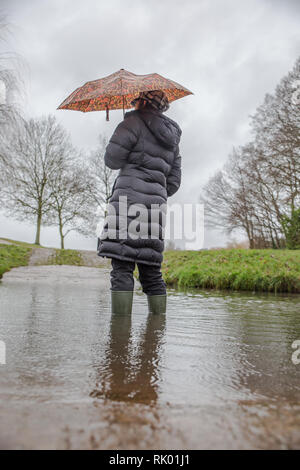 Kidderminster, Regno Unito. 8 febbraio, 2019. Regno Unito: meteo persistente pioggia pesante porta a livelli di acqua di risalita in Worcestershire con docce blustery continuando per tutto il giorno. Una giovane donna, vista posteriore, che indossa un lungo cappotto invernale e wellies sorge isolata all'esterno in acqua, lottando con il suo ombrello in umido e il vento. Credito: Lee Hudson/Alamy Live News Foto Stock