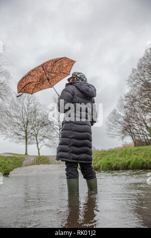 Kidderminster, Regno Unito. 8 febbraio, 2019. Regno Unito: meteo persistente pioggia pesante porta a livelli di acqua di risalita in Worcestershire con docce blustery continuando per tutto il giorno. Una giovane donna, vista posteriore, che indossa un lungo cappotto invernale e wellies sorge isolata all'esterno in acqua, lottando con il suo ombrello in umido e il vento. Credito: Lee Hudson/Alamy Live News Foto Stock