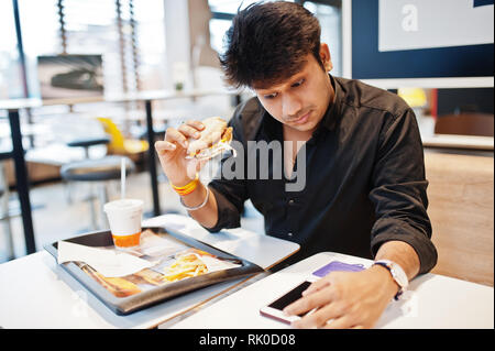 Indiano elegante uomo seduto al fast food cafe e mangiare hamburger, leggere morning news sul telefono cellulare e hanno sorpreso gli occhi. Foto Stock