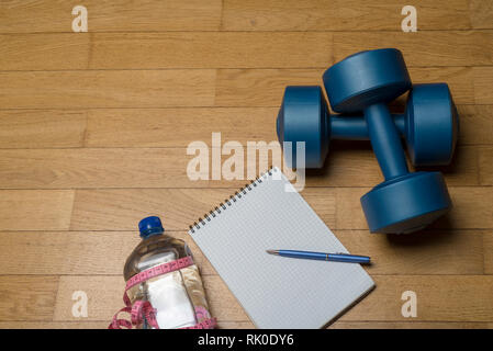 Formazione, esercizio, allegria e salute - due manubri in plastica, un notebook, acqua minerale e una penna su pavimento di legno. Il concetto di una sana Foto Stock