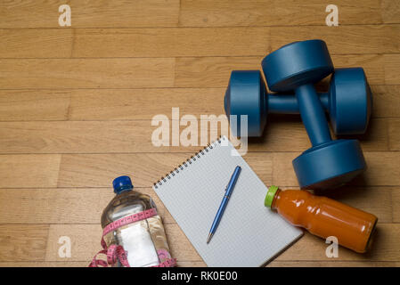 Formazione, esercizio, allegria e salute - due manubri in plastica, un notebook, acqua minerale con succo di frutta e una penna su pavimento di legno. Il concetto o Foto Stock