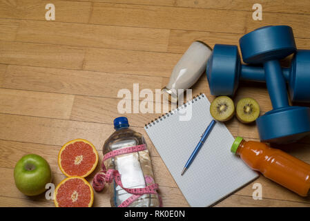 Formazione, esercizio, allegria e salute - due manubri in plastica, un notebook, acqua minerale con succo di frutta e una penna su pavimento di legno. Il co Foto Stock