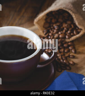 Primo piano di una fumante tazza di caffè con i chicchi di caffè versato da un sacchetto di tela in background Foto Stock