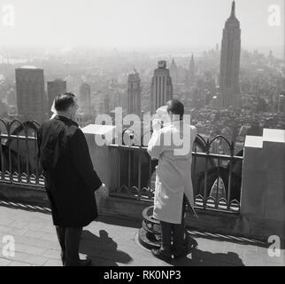 Degli anni Cinquanta, storico, guardando sopra la città di New York, New York, USA, due uomini in piedi in cima ad un alto edificio, uno utilizzando un visualizzatore o vista del telescopio, con l'Empire State edifico in distanza. Foto Stock