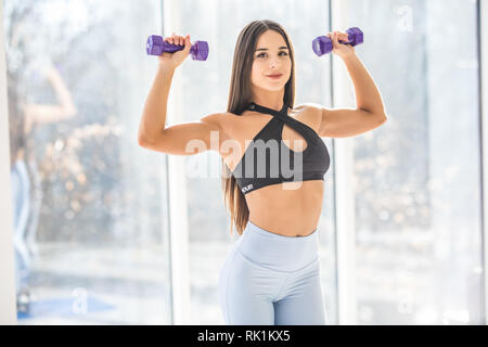 Atletica Giovane donna pompando i muscoli con manubri al fitness club di fronte a finestre. Foto Stock