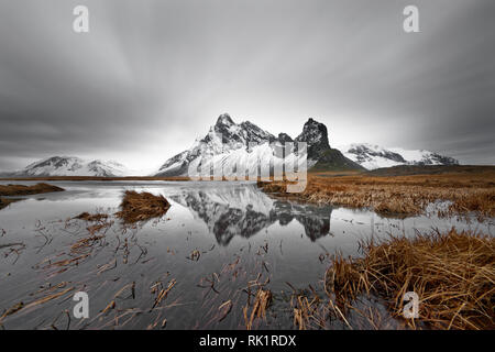 Coperto di neve e la formazione di montagna è riflessa in uno stagno, ciuffi di erba in e intorno allo stagno, lame di erba sono parzialmente spostata dal vento, cloud Foto Stock