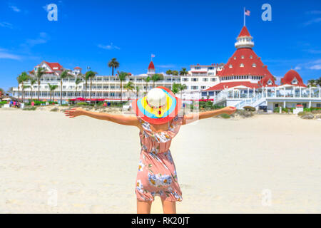 Carefree woman in hat su Coronado Central Beach, cercando storico hotel Vittoriano su Coronado Island, San Diego. Stile di vita godendo turistica estiva Foto Stock