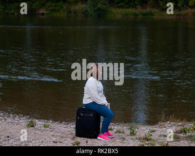 Un solitario triste giovane donna seduta attesa in una valigia sul bordo di un fiume Foto Stock