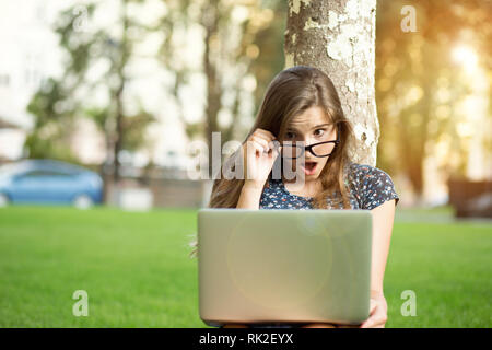 Scioccato adolescente guardando il computer tenendo bicchieri giù storditi all'aperto sdraiato sul prato verde le cuffie sulla testa. Il modello multiculturale di razza mista Foto Stock