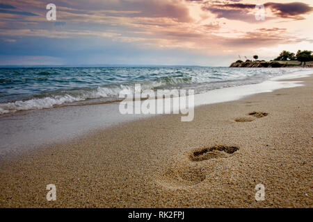 Bellissima vista sulla spiaggia di sabbia con impronte nella sabbia ondulata blu mare e cielo nuvoloso nel crepuscolo. Foto Stock