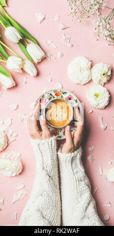 Flat-lay di mani femminili in maglia maglione holding tazza di caffè, bianco fresco e tulipani buttercup fiori su sfondo rosa, vista dall'alto. La molla vib Foto Stock