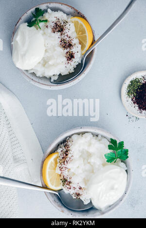 Appetitosa farinata di riso in due ciotole di ceramica guarnito con yogurt, sommacco, menta secca e spicchi di limone fotografato su sfondo grigio. Foto Stock