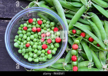 Sgusciate i piselli e fragole selvatiche in una ciotola di vetro e alcuni baccelli intorno sul vecchio tavolo in legno di quercia, vista da sopra Foto Stock