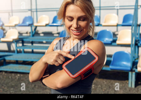 Donna giovane atleta che indossa fascia permanente sulla stadium stile di vita sportiva toccando lo schermo dello smartphone protector sorridente allegro close-up Foto Stock
