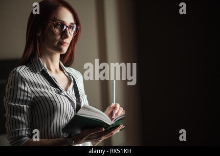 Lo zenzero intelligente giovane donna caucasica in seta camicetta a righe con sguardo premuroso, tenendo aperto il blocco note e penna la scrittura di note. Foto Stock