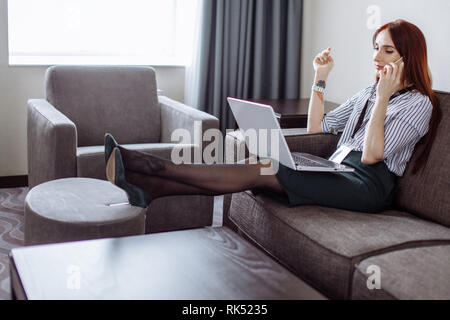Interessanti i capelli rossi femmina di project manager di marketing corporation indossando badge vuoto rendendo il lavoro a distanza seduta alla camera in hotel utilizzando laptop e Foto Stock
