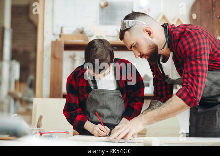 Laboriosa little boy è occupato facendo misurare e marcatura di asse di legno con una matita con suo padre a lavorare come carpentiere in seduta worksho in legno Foto Stock