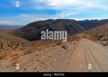 Deserto montagna strada sterrata che conduce verso la montagna sopra la valle sottostante sotto il luminoso cielo blu con nuvole bianche. Foto Stock