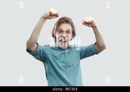 Ritratto di stupiti felice bello capelli lunghi biondo giovane uomo in blu T-shirt casual in piedi urlando e celebrare la sua vittoria. piscina studio shot Foto Stock