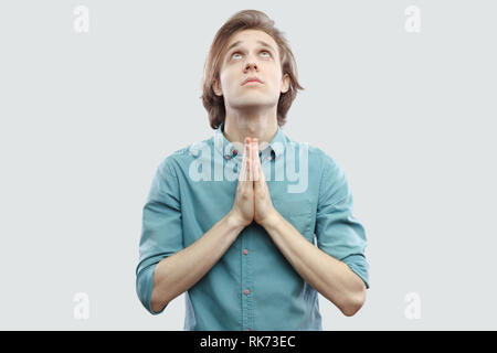 Ritratto di palm mani bello capelli lunghi biondo giovane uomo in blu T-shirt casual in piedi che guarda in alto e pregare o accattonaggio a Dio di perdonare lui. indoo Foto Stock