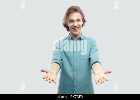 Ritratto di felice geniale bello capelli lunghi biondo giovane uomo in blu T-shirt casual in piedi con le braccia sollevate e sorriso toothy guardando la fotocamera. indo Foto Stock