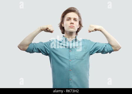 Io sono forte. Ritratto di orgogliosa bello capelli lunghi biondo giovane uomo in blu T-shirt casual in piedi e mostrando il suo bicipite e guardando la fotocamera. indoo Foto Stock