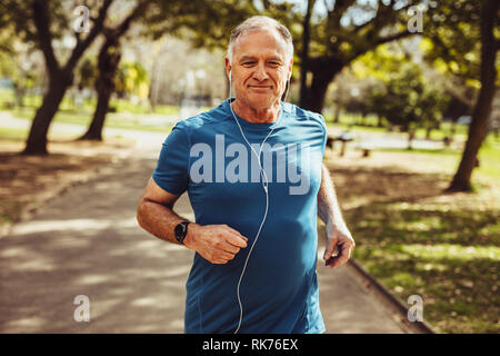 Allegro senior uomo a fare jogging nel parco ascoltando musica in cuffia. Senior persona fitness in esecuzione nel parco per una buona salute. Foto Stock