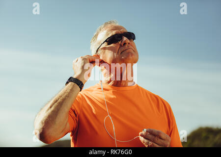 Ritratto di un senior athletic uomo in piedi all'aperto e ascoltare la musica tramite gli auricolari. Uomo in occhiali scuri mettendo su auricolari superarsi permanente Foto Stock