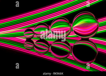 Quattro diverse dimensioni crystal / Vetro / lente di palline in una fila riflettendo come in uno specchio su un fondo nero con rosa e verde della luce al neon striature dietro Foto Stock