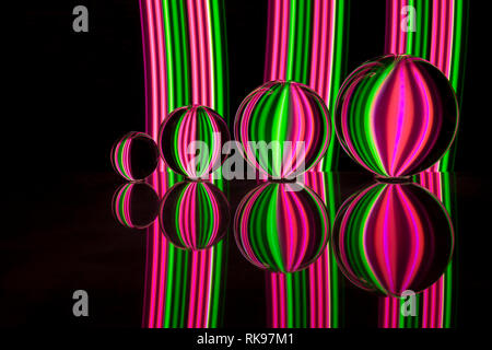 Quattro diverse dimensioni crystal / Vetro / lente di palline in una fila riflettendo come in uno specchio su un fondo nero con rosa e verde della luce al neon striature dietro Foto Stock