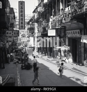 Anni '50, storico, diurno e una vista soleggiata di una strada nella città vecchia, Hong Kong, mostrando insegne con scrittura o caratteri cinesi e soggiorno con vestiti appesi, asciugando sui balconi sopra i negozi al dettaglio e venditori di strada al piano terra Foto Stock