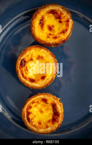 Posa piatto Immagine di tre Pastel de natas (PastÃ©è de BelÃ©m), su un blu piatto di terracotta portoghese di sfondo crema pasticcera crostate Foto Stock