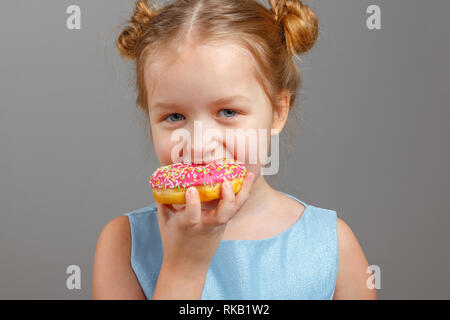 Un grazioso piccolo bambino ragazza in un abito blu morde una deliziosa ciambella rosa. Sfondo grigio, ritratto, studio. Foto Stock