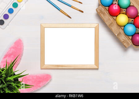 Un telaio, orecchie di coniglietto, un vaso di fiori artificiali con erba verde e colorati di uova in una scatola di cartone, vernici e pennelli. Concetto di pasqua. Spazio di copia Foto Stock