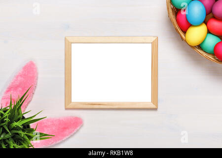 Telaio, orecchie di coniglietto, un vaso di fiori artificiali con erba verde, colorate le uova nel paniere. Concetto di pasqua. Spazio di copia Foto Stock