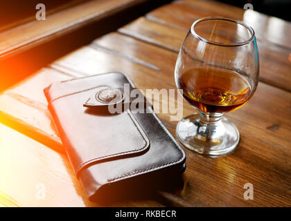 Bicchiere di whiskey o altro forte amber bevanda alcolica e pelle marrone portamonete sul nero tabella riflettente top studio sfondo, immagine orizzontale Foto Stock