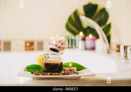 Caffè miscela scrub in un barattolo di vetro per l'uso di molti scopi: il viso, i capelli, maschera corpo e scrub concetto, al chiuso nel bagno di casa, candele accese fiamme sul th Foto Stock