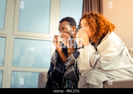 Giovane seduto vicino alla finestra nel soggiorno avente starnuti Foto Stock